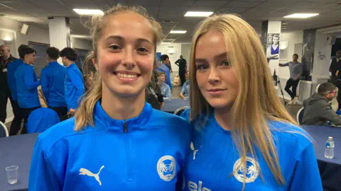 Tom Williams/BBC Jess Abrantes and Katie Gee smiling at the camera wearing blue Peterborough United strips. Other players in blue strips can be seen behind them.