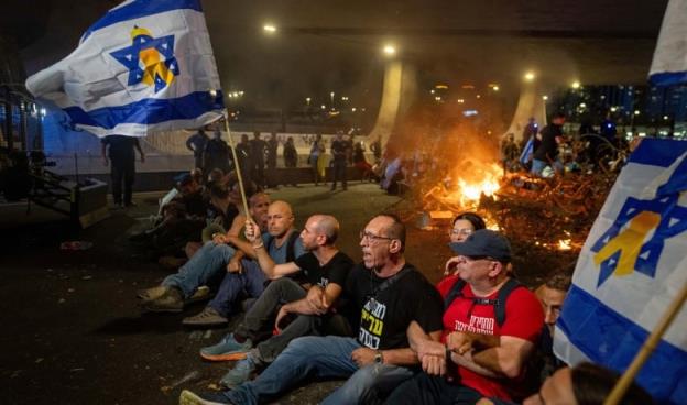 A line of men sit on the ground with linked arms, some holding Israeli flags. A fire burns in the road behind them. 